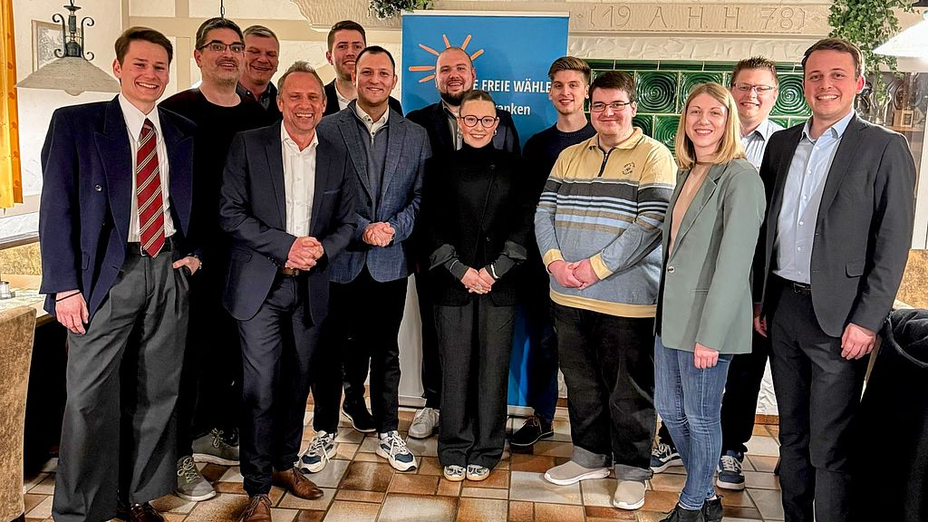 Eine Gruppe von 13 Personen unterschiedlichen Alters steht lächelnd in einem rustikalen Innenraum für ein Gruppenfoto zusammen. Im Hintergrund sind teilweise ein Roll-up-Banner der Partei sowie ein markanter, grüner Kachelofen zu erkennen.