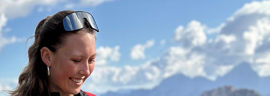 Lena Fischer steht lächelnd in dunkler Sportkleidung und mit einem roten Rucksack auf einem steinigen Bergpfad. Im Hintergrund erstreckt sich ein weites Bergpanorama unter einem blau-weißen Himmel.